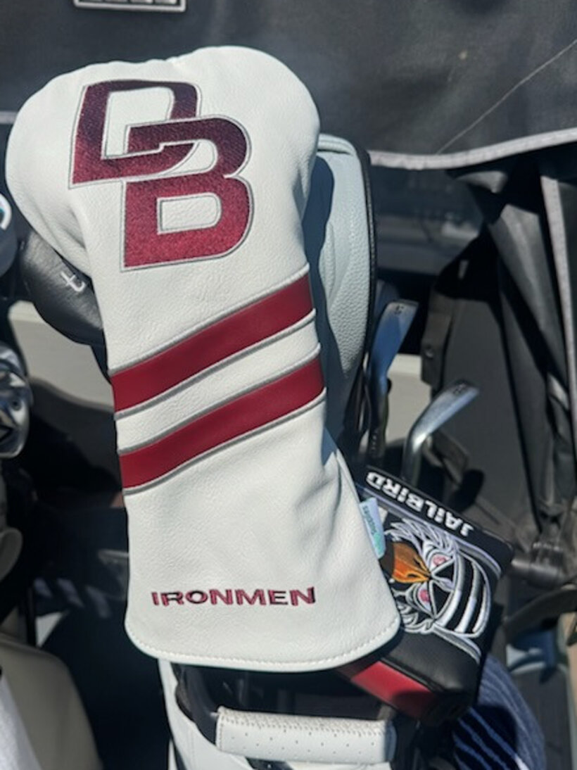 A white driver headcover with a maroon DB monogram and IRONMEN, in a golf bag on a cart
